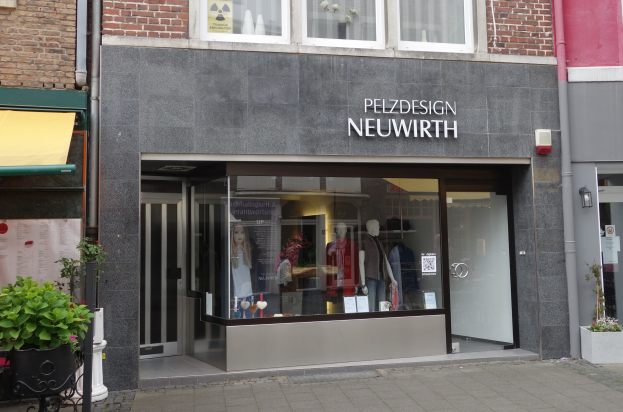 Eine Schaufensterfront mit Glaswänden und -türen, die Puppen in Kleidern zeigen, Text an der Wand, Topfpflanzen und ein Schild mit der Aufschrift "Pelzdesign Neuwirth in Amsterdam".