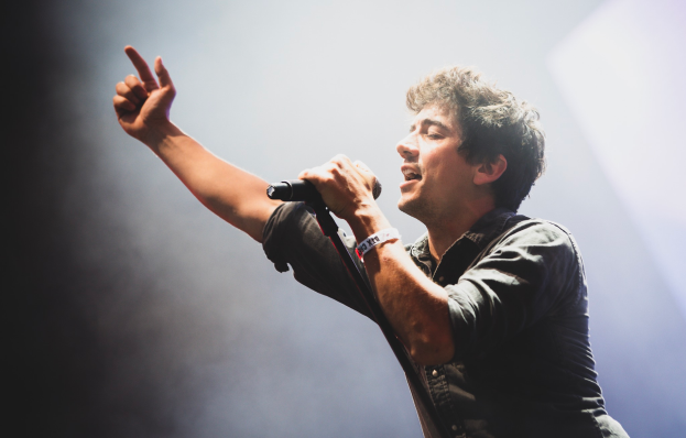 Ein Mann in schwarzem Hemd und Jeans singt leidenschaftlich in ein Mikrofon auf einer Bühne, mit hellen Scheinwerfern und einem jubelnden Publikum im Hintergrund.