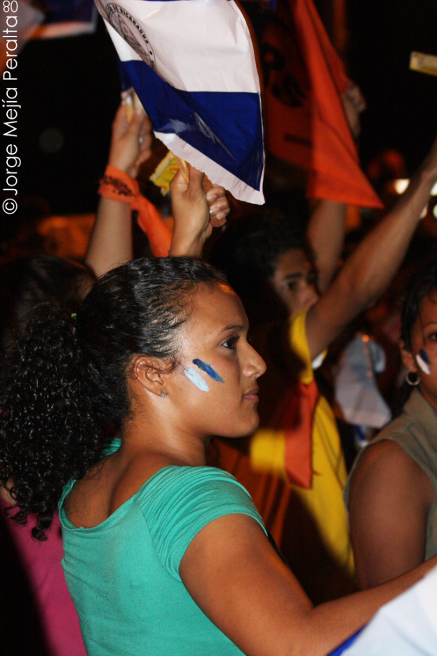 Eine Frau in einem grünen T-Shirt steht bei einer Demonstration und hölt ein weißes Schild, mit vielen anderen Menschen hinter ihr.