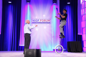 Eine Person steht auf der Bühne und spricht, während eine andere Person auf einem Fahrzeug hinter ihnen steht, vor blauen Vorhängen, Scheinwerfern und einem Banner.