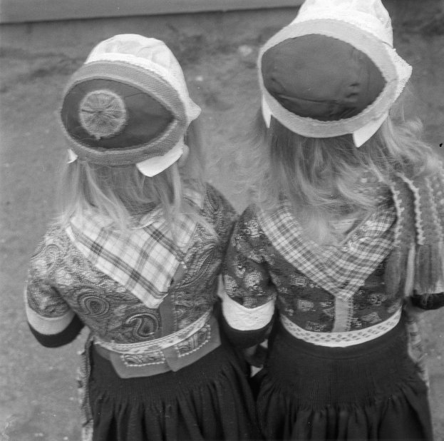 Zwei junge Mädchen in traditionellen bayrischen Trachten und Hüten stehen nebeneinander vor einer Wand und werden in Schwarz-Weiß abgebildet.