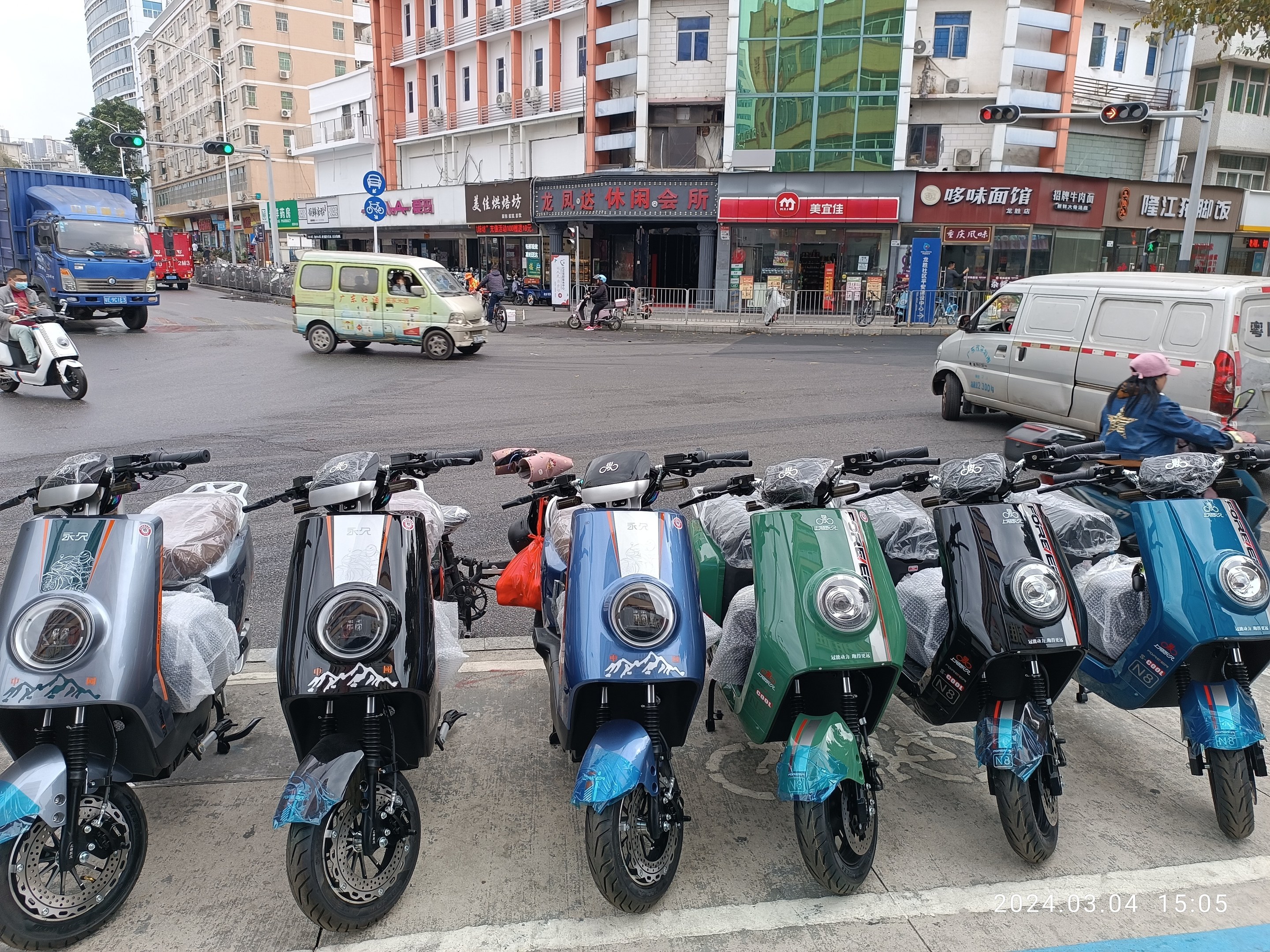 Eine Reihe von elektrischen Rollern, die auf einer Straße geparkt sind, mit einer Person in einer Mütze davor, vor Häusern, Bäumen und einem klaren blauen Himmel.