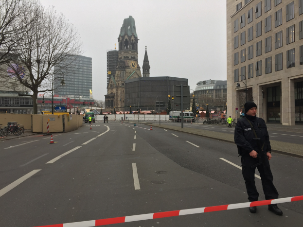 Ein Polizist in schwarzer Uniform und Mütze steht in einer Berliner Straße umgeben von Verkehrskegeln, Fahrrädern, Laternen, Bäumen und Gebäuden, mit einem Kirchturm im Hintergrund unter einem klaren blauen Himmel.