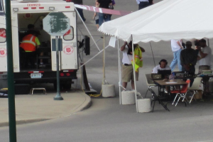 Ein Krankenwagen steht links von einer Straße, während rechts ein Zelt viele Menschen schützt, mit einem Tisch und einem Stuhl in der Nähe.