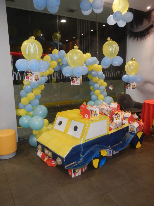 Eine McDonald's-Geburtstagsfeier mit einem gelben und blauen Lastwagen, der mit Ballons und anderen Dekorationen geschmückt ist, einem Tisch mit roter Decke und Gegenständen, Hockern, einer Glaswand und Deckenlampen in einer festlichen Atmosphäre.