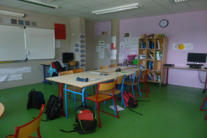 Ein Klassenzimmer mit Tischen, Stühlen, Taschen, Bücherregalen, einem Monitor, Tastatur, Maus, Uhr, Papieren an den Wänden, einer Tafel und Deckenleuchten.