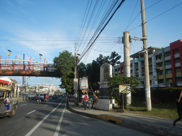 Eine belebte Stadtstraße mit fahrenden Fahrzeugen, Fußgängern auf dem Gehweg, Strommasten mit Drähten, Bäumen, mehrstöckigen Gebäuden mit Fenstern, einer Brücke mit Geländern im Hintergrund, Straßenlaternen und einem bewölkten Himmel.
