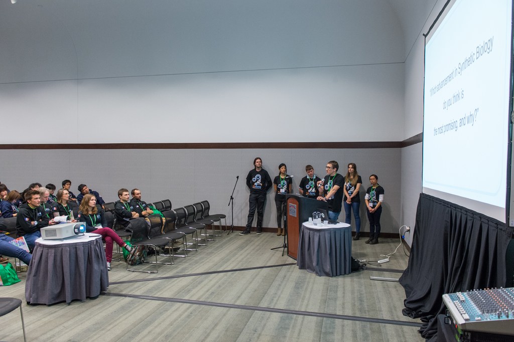 Eine Person, die in ein Mikrofon spricht, bei einer Versammlung mit einem Publikum, Teamkollegen mit ID-Karten, ein Präsentationsbildschirm und ein Videoabspieler in einem leeren Büroraum.