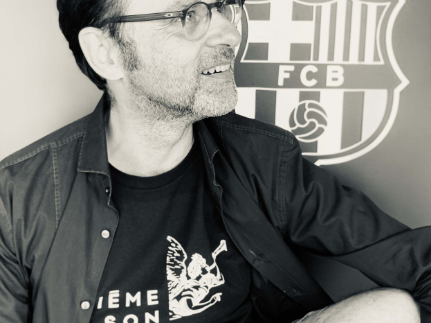 Ein Mann in Brille und schwarzem Hemd lächelt vor einem schwarz-weißen FC Barcelona-Logo an einer Wand.