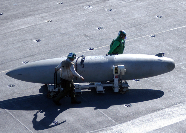 Zwei Männer in Schutzausrüstung inspizieren eine große, spitz zulaufende zylindrische Rakete, die wie eine Atombombe aussieht, auf einem Flugzeugträger.